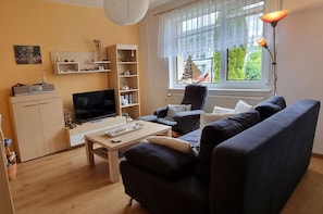 A living room with a sofa, TV, shelves, and a window with a view. - Vacation apartment Kollwitz - very close to the Sassnitz town harbor - Holiday apartment (Sassnitz auf Rügen)