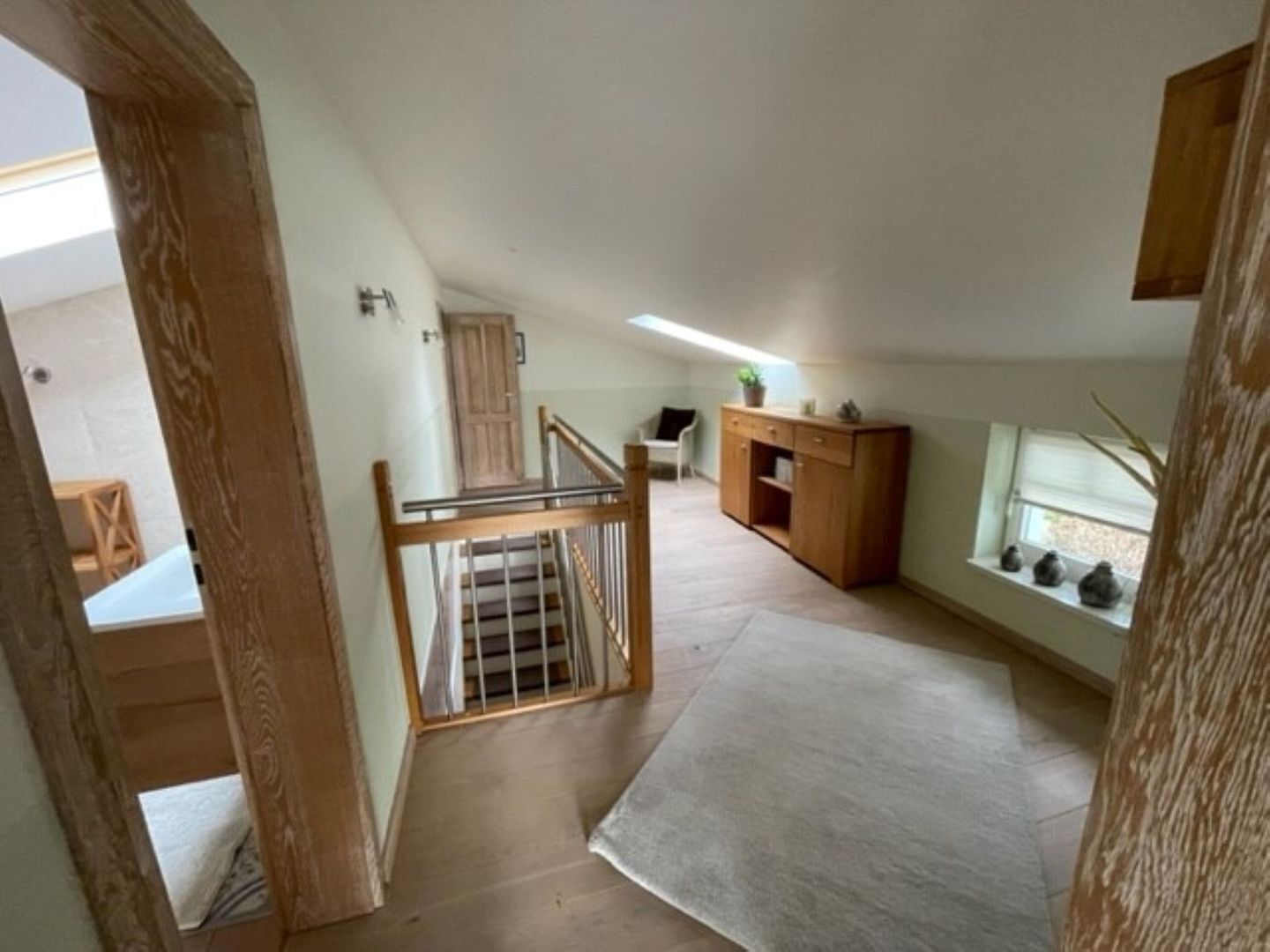 A room with a wooden staircase, a desk, a window with plants, and a door leading to another room.