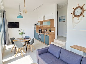 A modern kitchen with wooden cabinets, a dining area with a table and chairs, a sofa, and a wall-mounted clock. - Villa Küstenperle, App. Muschelblick - pure sea view! (Sassnitz auf Rügen)