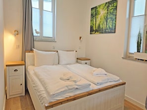 A bedroom with a bed, bedside tables, a nightstand, a window with blinds, a wall-mounted light, and a painting. - Villa Küstenperle, App. Muschelblick - pure sea view! (Sassnitz auf Rügen)