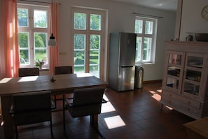 A kitchen with a dining table, chairs, a refrigerator, a trash bin, and a cabinet with glass doors. - Jasmundhus on Rügen - Holiday apartment Backbord (Neddesitz auf Rügen)