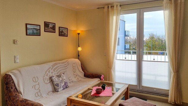 Ein gemütliches Zimmer mit einer Couch, einem Glastisch, einem kleinen Tisch mit Platzdeckchen und einem Fenster mit Vorhängen.