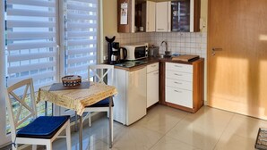 Eine kleine Küche mit Esstisch, weißen Schränken, einer Kaffeemaschine und einem Fenster mit Jalousien.