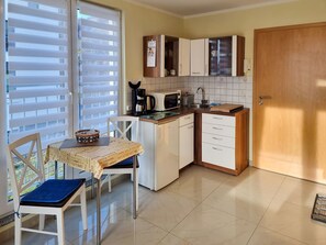 Eine kleine Küche mit Esstisch, weißen Schränken, einer Kaffeemaschine und einem Fenster mit Jalousien.