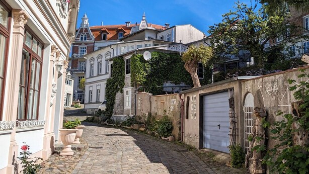 Una calle adoquinada flanqueada por edificios, incluyendo un edificio blanco con balcón y otro de techo rojo al fondo.