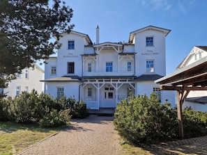 Ein zweistöckiges Gebäude mit einem zentralen Eingang, beschriftet als 'Villa Bertha's Heim', umgeben von Grünpflanzen und einem Pflasterweg.