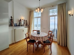 Ein Esszimmer mit rundem Tisch, Holzstühlen, einer Chandelüster, einer Heizung und Blick durch große Fenster.