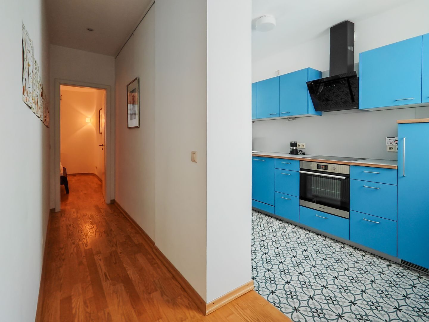 A hallway leading to a kitchen with blue cabinets, a black range hood, and patterned floor tiles.