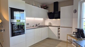A modern kitchen with white cabinetry, a built-in oven, a coffee machine, and a window providing natural light. - Villa Meerzeit - Holiday apartment Mare Azur (Sassnitz)
