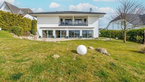 A modern two-story house with a large glass front, a balcony, and a well-maintained lawn with a spherical sculpture. - Villa Meerzeit - Holiday apartment Mare Azur (Sassnitz)