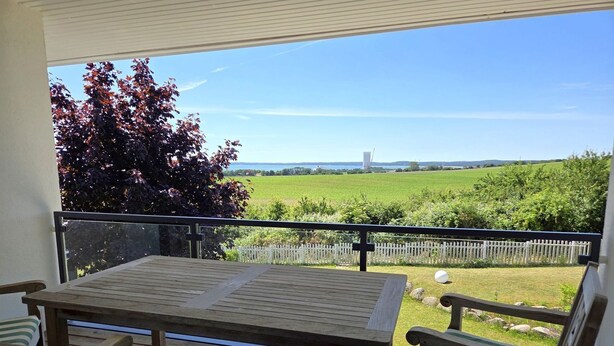 A wooden deck with a table and chairs, a white picket fence, and a view of a green field and distant trees. - Villa Meerzeit - Holiday apartment Mare Azur (Sassnitz)