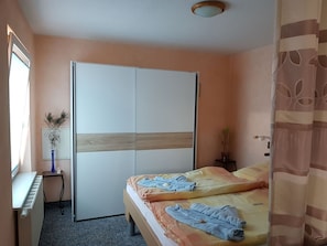 Ein Schlafzimmer mit Bett, Schrank, Heizkörper, einem kleinen Tisch mit Vase und einer Topfpflanze.