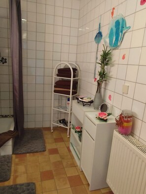A bathroom with a white tiled wall, a white radiator, a white shelf unit, and a towel rack with folded towels. - Dornbusch vacation apartment - quiet location on the outskirts - Dornbusch vacation apartment - in (Sassnitz auf Rügen)
