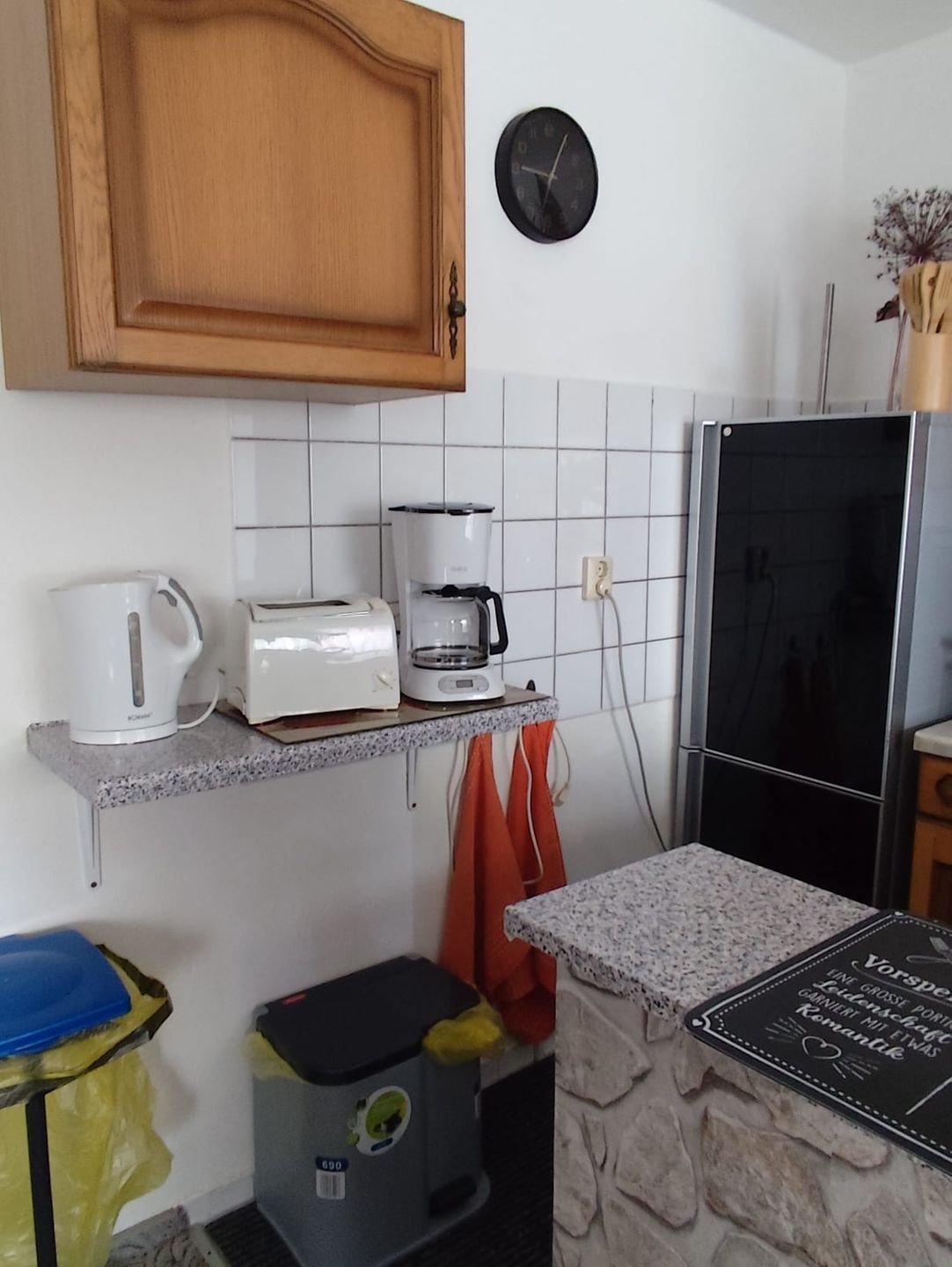 A kitchen with a wooden cabinet, a coffee maker, a toaster, a clock, a refrigerator, and a trash bin.