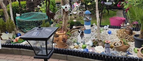Puutarha, jossa on monenlaisia kasveja, lintujen ruokintalaite ja koristeellinen aita.