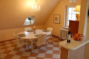 Un coin repas avec un sol à carreaux, une table ronde, des chaises blanches, un lustre, un radiateur, une fenêtre donnant sur un beau paysage et un saladier rempli d’oranges posé sur le comptoir.