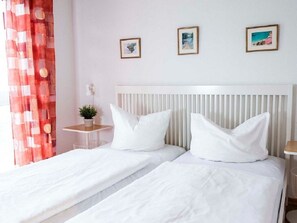 Uma cama de casal com roupa de cama branca, uma mesinha de cabeceira com uma planta e duas fotos emolduradas na parede.