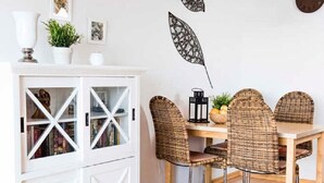 Un comedor con un armario blanco, sillas de mimbre, una mesa de madera y hojas decorativas en la pared.