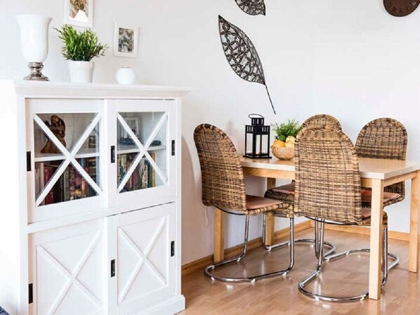 Uma área de refeições com um armário branco, cadeiras de vime, uma mesa de madeira e folhas decorativas na parede.