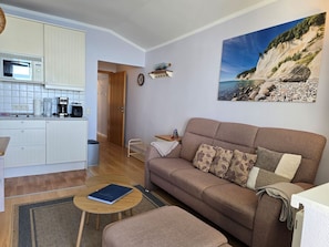 Ein Wohnzimmer mit einer Couch, einem Couchtisch, einer Küche mit weißen Schränken, einer Mikrowelle und einem Bild von einer Küstenlandschaft.