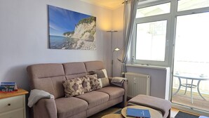 A living room with a sofa, a small table, a lamp, and a painting of a coastal scene. - Fürstenhof App. 405 - with balcony and sea view - Fürstennhof, App. 405 - 1. (Sassnitz auf Rügen)