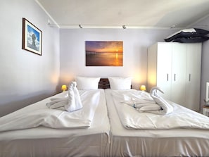 Ein Hotelzimmer mit zwei Betten, einem Abendsonnen-Gemälde, einem Kleiderschrank und einer Lampe auf dem Nachttisch.