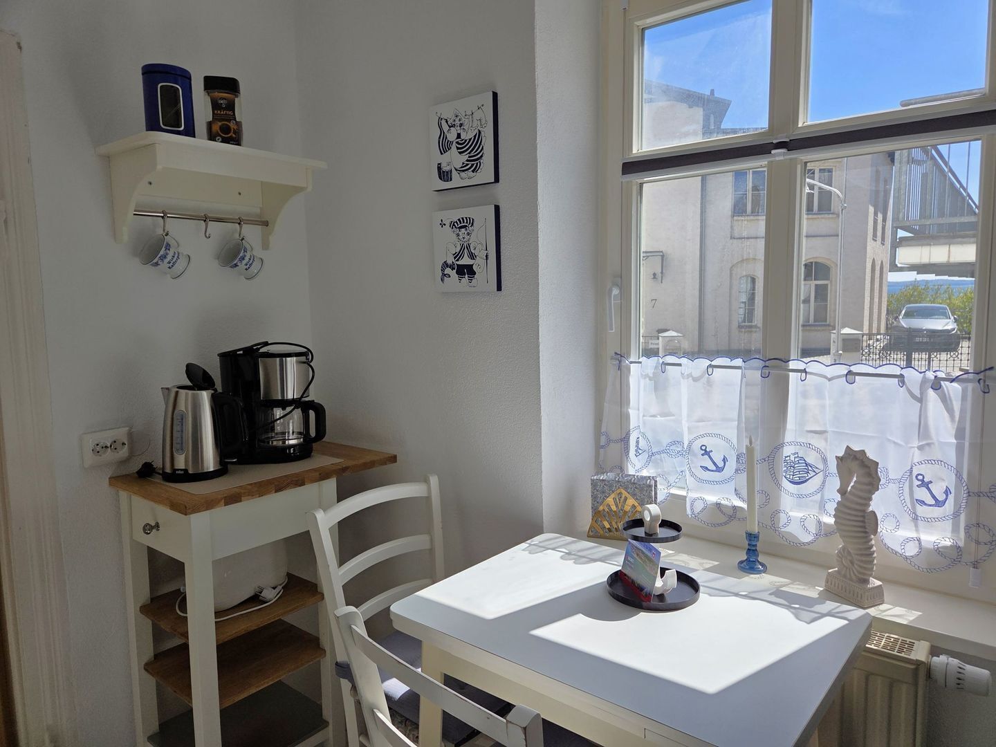 Une petite cuisine équipée d’une machine à café, d’une table blanche et d’une fenêtre donnant sur les bâtiments environnants.