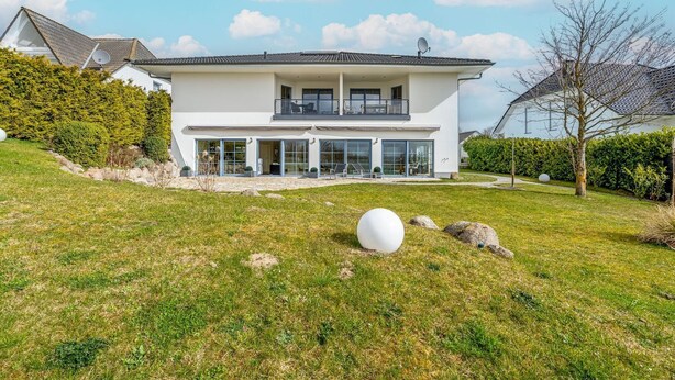 Une maison moderne à deux étages, dotée d’une grande façade vitrée, d’un balcon et d’une pelouse bien entretenue agrémentée d’une sculpture sphérique.
