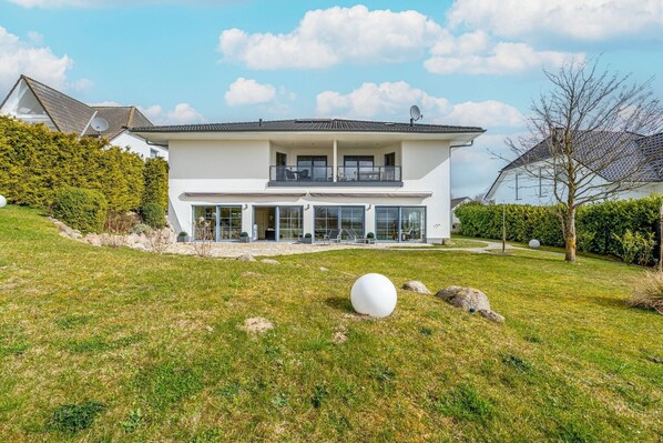 Ein modernes, zweistöckiges Haus mit großer Glasfront, einem Balkon und einem gepflegten Rasen mit einer kugelförmigen Skulptur.