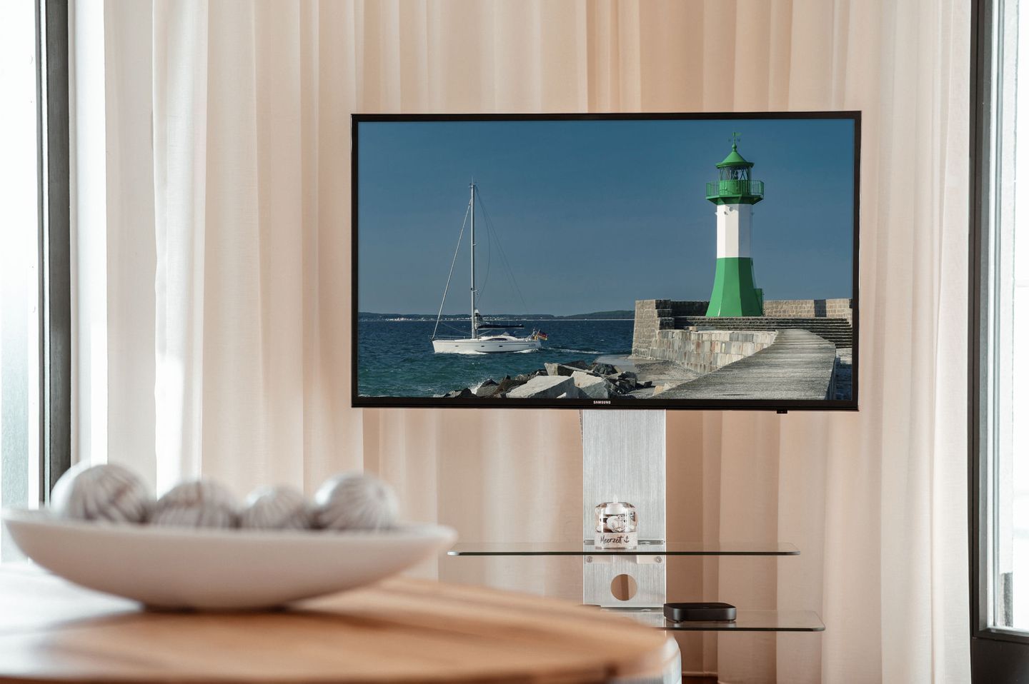 Ein an der Wand befestigter Fernseher zeigt eine Küstenansicht, auf einem Tisch steht eine Obstschale und das Samsung-Logo ist zu sehen.