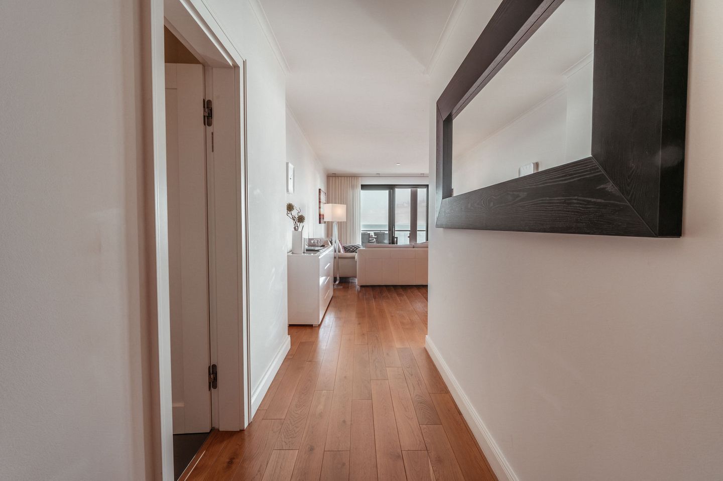 Een hal met houten vloer, een witte deur, een witte bank, een tafel en een raam met uitzicht.