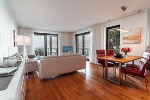 A modern living room with a sofa, dining table, and chairs, large windows with curtains, and a flat-screen TV. - Villa Friede-Marie, Apt. Freya - with a magnificent view of the sea - Villa Friede-Marie, (Sassnitz auf Rügen)