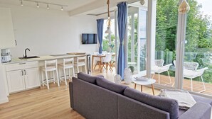 Una cocina moderna con armarios blancos, un comedor con sillas blancas, un sofá gris y un balcón con sillas y una mesa blancos.