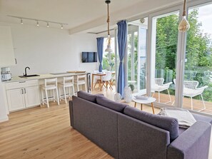 Eine moderne Küche mit weißen Schränken, ein Essbereich mit weißen Stühlen, ein graues Sofa und ein Balkon mit weißen Stühlen und einem Tisch.