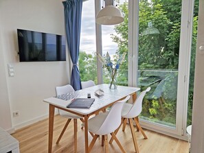 Ein stilvoller Essbereich mit Tisch, Stühlen, einer Blumenvase, einem an der Wand montierten Fernseher und einem großen Fenster mit Blick auf Grünflächen.