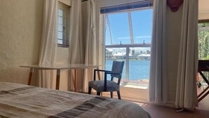 Ein Schlafzimmer mit Bett, Stuhl, Tisch und einem großen Fenster mit Blick auf ein Gewässer.