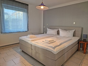 Ein Doppelbett mit Kopfteil, ein Nachttisch mit Lampe und ein Fenster mit Vorhängen.