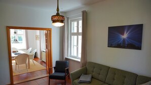 Ett vardagsrum med en grön soffa, ett matbord i trä med stolar, en mönstrad matta, en hängande lampa och en tavla som föreställer en fyr.