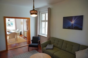Ein Wohnzimmer mit einem grünen Sofa, einem Holzespeisesaal mit Stühlen, einem gemusterten Teppich, einer Hängeleuchte und einem Gemälde eines Leuchtturms.