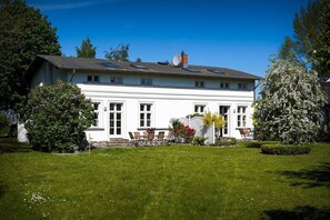 Ein weißes, zweistöckiges Haus mit Flachdach, Schornstein und einem Garten mit Rasen, einer Bank und einem Baum.