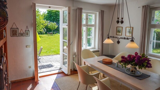 En öppen dörr som leder ut till en trädgård, ett matbord med stolar och en hängande lampa.