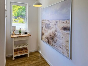 Ein Zimmer mit Fenster, einem Schreibtisch mit Lampe, einem Lautsprecher und einem großen Gemälde mit Strandmotiv.