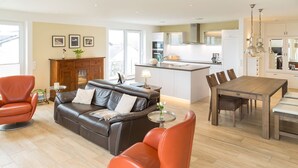 A modern living room with a leather sofa, orange armchairs, a dining table, and a kitchen in the background. - Villa am Kreidebruch, FeWo OstseeWeitblick - panoramic view of the Baltic Sea (Sassnitz auf Rügen)
