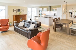 A modern living room with a leather sofa, orange armchairs, a dining table, and a kitchen in the background. - Villa am Kreidebruch, FeWo OstseeWeitblick - panoramic view of the Baltic Sea (Sassnitz auf Rügen)