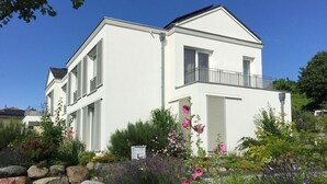 A two-story white building with shutters, a balcony, and a garden with various plants and flowers. - Apartment Richi - central and quiet in the harbor town of Sassnitz - (Sassnitz auf Rügen)