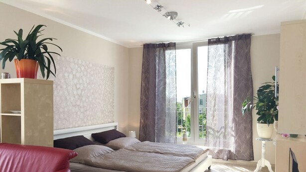 A bedroom with a bed, a red chair, a bedside table with a lamp, and a window with curtains. - Apartment Richi - central and quiet in the harbor town of Sassnitz - (Sassnitz auf Rügen)