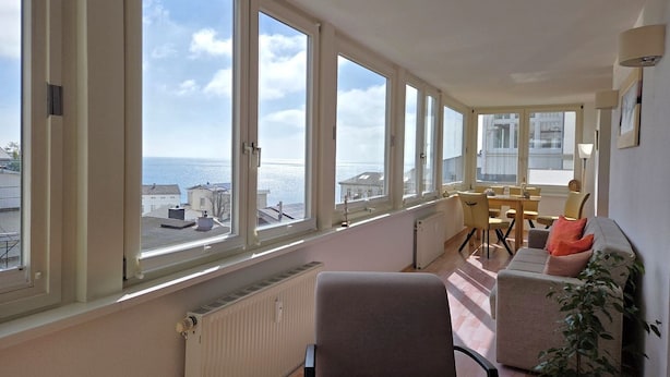 Um quarto espaçoso com grandes janelas que oferecem vista para o mar e edifícios, uma área de refeições com cadeiras e mesa, e um radiador ao longo da parede.