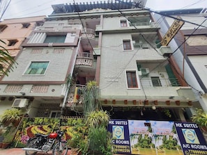 Ein mehrstöckiges Gebäude mit Balkonen, ein Motorrad davor sowie Schilder mit den Aufschriften 'MK SUITES' und 'A/C ROOMS'.