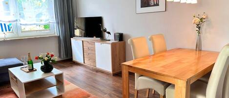 A modern living room with a wooden dining table, white chairs, a TV, a bookshelf, and a window with curtains.