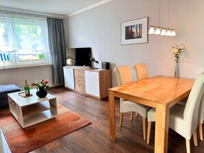 A modern living room with a wooden dining table, white chairs, a TV, a bookshelf, and a window with curtains. - Vacation apartment Seestern - Holiday apartment Seestern in Sassnitz on Rügen with (Sassnitz auf Rügen)